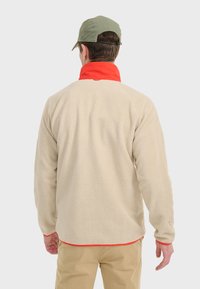 Veste en polaire beige avec une surface texturée et un col rouge, dotée de manches longues et d'une coupe décontractée. Complétée par une casquette verte.