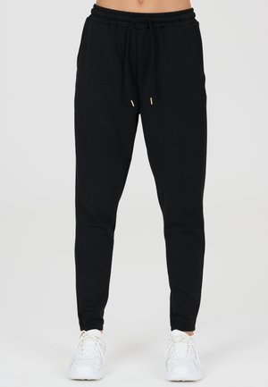 Person trägt schwarze Jogginghose mit Kordelzug in der Taille und weiße Sportschuhe, steht vor einem einfachen hellen Hintergrund.