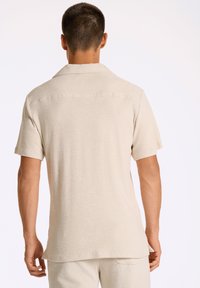 Camicia beige a maniche corte realizzata in tessuto morbido e dalla texture piacevole, con colletto e orli dritti, caratterizzata da un sottile dettaglio della cucitura posteriore.