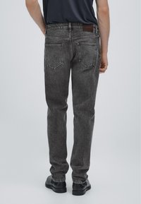 Bakifrån av en person som bär svarta acid-wash jeans, svarta skor och en instoppad svart skjorta mot en enkel bakgrund.