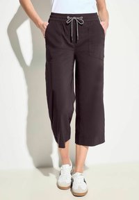 Schwarze, kürzere Hosen mit einem Kordelzugbund, ausgestattet mit zwei Fronttaschen und einer glatten Textur. Kombiniert mit weißen Sportschuhen.