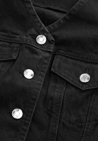Schwarze Jeansjacke mit silbernen Druckknöpfen, einem V-Ausschnitt-Kragen und zwei Brusttaschen mit umgeschlagenen Klappen. Sichtbare Strukturstoffdetails.