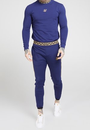 Chándal deportivo en color marino, hecho de tejido elástico; parte superior de manga larga con un logo, pantalones con franjas laterales y un cinturón estampado, combinados con zapatillas blancas.