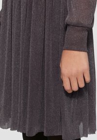 Graues plissiertes Kleid mit strukturiertem Stoff, der leicht schimmert. Lange Ärmel mit gerippten Bündchen. Hand ruht am Stoffrand.