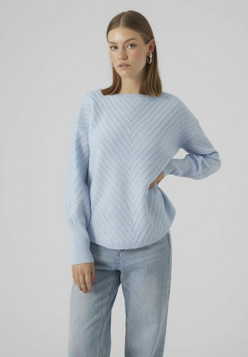 Svetlo modra pletena pulover s chevron vzorcem, širokim čolničastim izrezom in rebrastimi manšetami. V kombinaciji s svetlimi kavbojkami.
