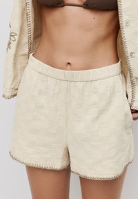Shorts bej dintr-un material texturat. Au o talie elastică și cusături decorative de-a lungul tivurilor pentru un plus de detaliu.