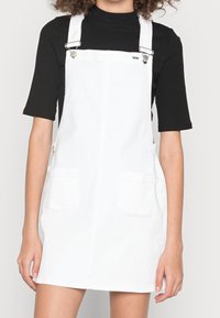 Vit denim overallklänning med två framfickor, justerbara metallremmar och rak skärning. Texturen ser slät och lätt ut.