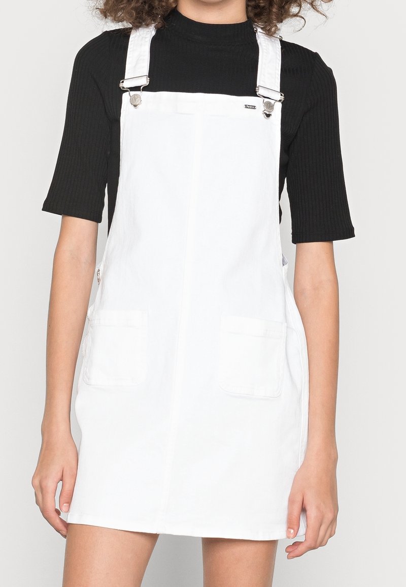 Vit denim overallklänning med två framfickor, justerbara metallremmar och rak skärning. Texturen ser slät och lätt ut.