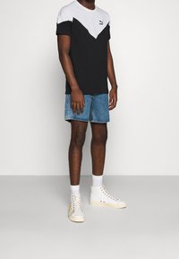 Muž v černobílém tričku Puma, modrých džínových šortkách, bílých ponožkách a vysokých bílých teniskách stojící na šedé podlaze.