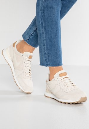 Osoba w jasnobeżowych sneakersach z białymi podeszwami i karmelowymi detalami, zestawionych z niebieskimi skróconymi dżinsami na jasnym tle.