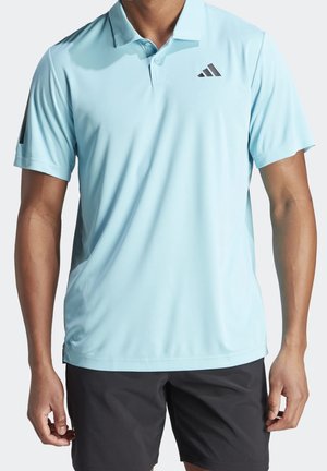 Homme portant un polo bleu clair à manches courtes avec col et patte de boutonnage, et un short noir, debout devant un fond uni.