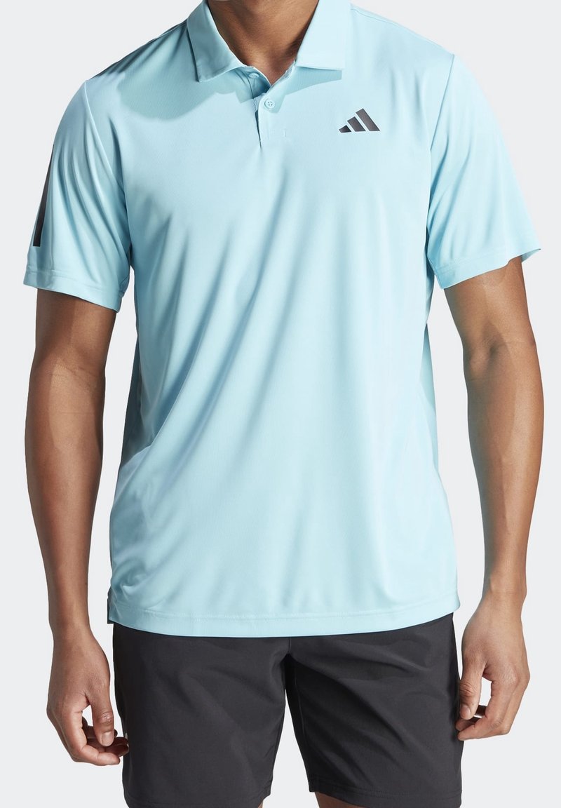 Homme portant un polo bleu clair à manches courtes avec col et patte de boutonnage, et un short noir, debout devant un fond uni.