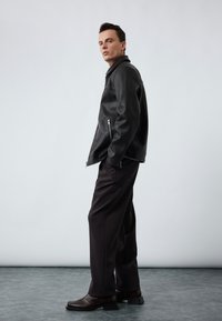 Zwarte leren jas met een staande kraag en ritszakken, gecombineerd met donkere broek en bruine leren schoenen. Gladde textuur, klassiek ontwerp.