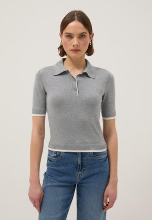 Mujer joven vistiendo una camiseta tipo polo ajustada de manga corta gris con ribete blanco y vaqueros azules, posando contra un fondo liso.