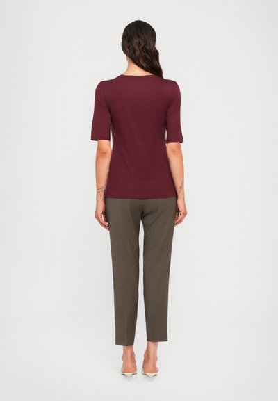 Woman with long dark hair stands facing away, wearing a burgundy short-sleeve top, gray pants, and beige low-heeled shoes.