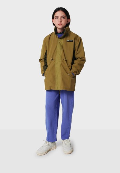 Olivgrön jacka med hög krage, framknappar och fickor; kombinerad med blå byxor och vita sneakers. Slät tygtextur.