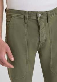 Pantalons en coton vert olive avec une fermeture à bouton, deux poches avant et un détail de couture prononcé. Texture lisse, coupe ajustée, design décontracté.