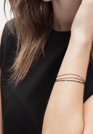 Bras gauche d'une personne à la peau claire et aux cheveux bruns portant un haut noir et un délicat bracelet noir à multiples chaînes fines au poignet.