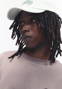Casquette de baseball blanche avec du texte vert, portée avec un sweat-shirt beige clair. Le sujet a des dreadlocks et une expression neutre.