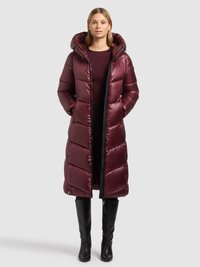 Bordowy pikowany długi płaszcz z wysokim kołnierzem i kapturem, zapinany na zamek błyskawiczny, z poziomym pikowaniem, noszony na czarnej bluzce i spodniach.