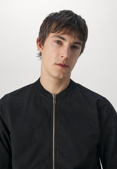 Chaqueta bomber negra con cuello acanalado, cierre frontal de cremallera y textura suave. No presenta patrones ni acentos visibles.