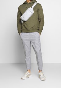 Olivgrön hoodie, ljusgrå rutiga byxor och vita sneakers med gula accenter. Grå axelväska med dragkedjestängning och logotyp.