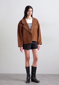 Giacca oversize in suede marrone con rever a notched, chiusura a bottoni, indossata sopra una camicia a righe bianca e nera, abbinata a pantaloni corti neri e stivali fino al ginocchio.