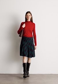 Κόκκινο μακρυμάνικο πουλόβερ με λαιμόκοψη, πράσινη καρό πλισέ φούστα, μαύρες μπότες μέχρι το γόνατο και καφέ τσάντα. Λείες υφές και τολμηρά σχέδια.