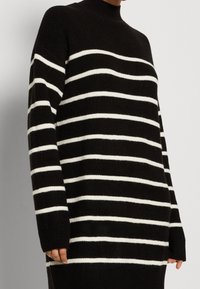 Maglione a collo alto a righe bianche e nere, con una texture a coste, maniche lunghe, vestibilità ampia, strisce bianche prominenti su sfondo nero.