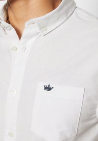 Vit knäppskjorta i bomull, med klassisk krage, vänster bröstfickan och blå kungakrona broderi på fickan.