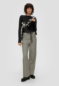 Schwarzes Langarmoberteil, karierte weite Hosen mit Kordelzugtaille und klobigen schwarzen Plateau-Schuhen. Pullover über die Schultern gewickelt.