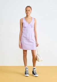 Abito senza maniche color lilla con scollo a V, corpino aderente e pieghe sottili. Abbinato a sneaker alte nere e una borsa bianca.