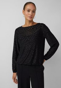 Vrouw met haar vastgebonden, gekleed in een zwart, lang mouwenshirt met een textuur en subtiele dierenprint, en zwarte broek, staand met één hand op de heup.