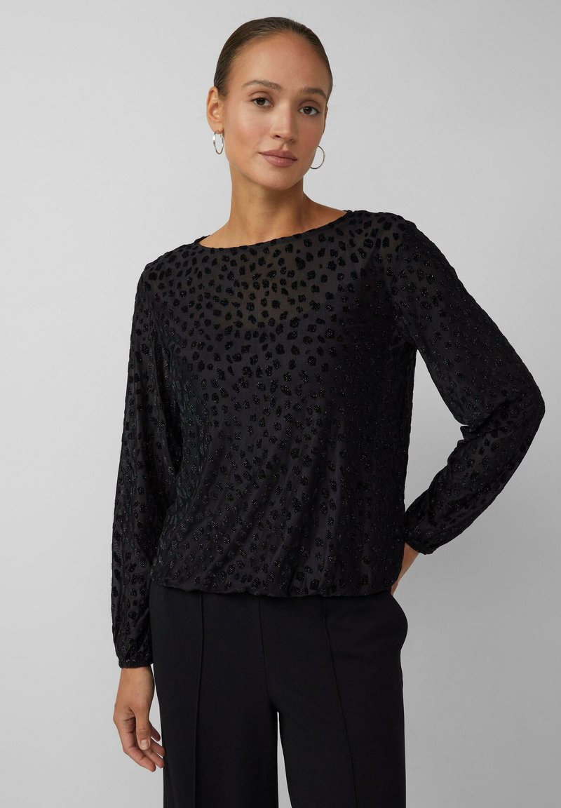 Vrouw met haar vastgebonden, gekleed in een zwart, lang mouwenshirt met een textuur en subtiele dierenprint, en zwarte broek, staand met één hand op de heup.