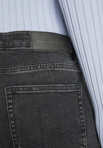 Jeans de ganga pretos com uma cintura texturizada, apresentando um rótulo de couro com a inscrição "Amanhã". Vista em close que destaca os detalhes da costura.