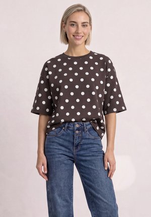 Ung kvinde smiler, iført en sort oversized t-shirt med hvide polkaprikker og blå high-waist jeans, står mod en ensfarvet baggrund.