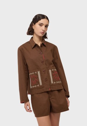 Femme portant une veste marron boutonnée avec des motifs floraux brodés sur les poches avant et un short marron assorti, debout devant un arrière-plan uni.