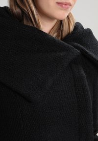 Woman with blonde hair wearing a textured black wool coat with a wide, folded collar, showing her lower face and neck.