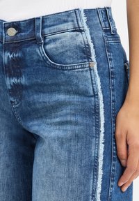 Jean en denim avec des dégradés de lavage clair et foncé, présentant une couture latérale effilochée et des rivets en métal, portés avec un haut blanc.