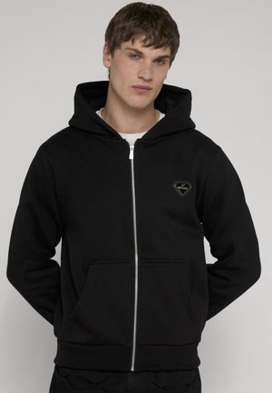 Jonge man met kort bruin haar die een zwarte hoodie met rits draagt met een klein logo op de borst, staand tegen een effen achtergrond.
