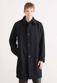 Cappotto in lana nera con colletto, bottoni frontali e tasche laterali. Presenta una linea dritta e un tessuto testurizzato.