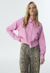 Roze overhemd met kraag en een zak met daarop de tekst "Diamond Lights", gecombineerd met hoog-was leeuwenhuid jeans. Beide hebben een ontspannen fit.