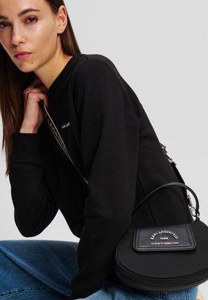 Sac à main circulaire en cuir noir avec bandoulière amovible, présentant un logo gravé à l'avant et une texture lisse. Porté de manière décontractée sur un haut sombre.