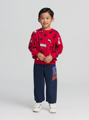 Ragazzo giovane in piedi che sorride, indossa una felpa rossa Marvel con grafiche di Spider-Man e pantaloni blu scuro con stampa di Spider-Man.