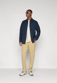 Camicia navy a bottoni sopra una maglietta crema, pantaloni beige e scarpe da ginnastica grigio chiaro con accenti neri. Modello che indossa occhiali da sole.