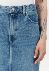 Denim sijonas su aukštu liemeniu, šviesiai mėlynos spalvos, priekinė kišenė ir metalinė sagčių detalė liemenyje.