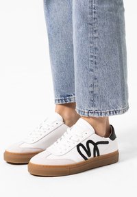 White sneakers with black wave design, padded tongue, and gum sole. Worn with light blue cuffed denim jeans. Smooth leather and textured accents.