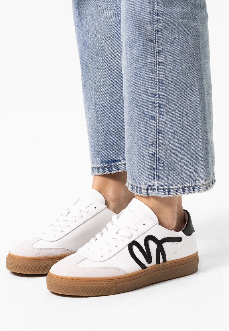 White sneakers with black wave design, padded tongue, and gum sole. Worn with light blue cuffed denim jeans. Smooth leather and textured accents.