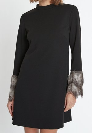 Zwarte jurk met lange mouwen, hoge halslijn en metallic franjes aan de manchetten, gedragen door een persoon van schouders tot halverwege de dij.