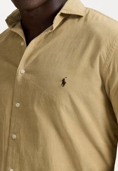 Close-up de uma pessoa vestindo uma camisa bege com botões e um logótipo de jogador de polo bordado em castanho escuro no peito.
